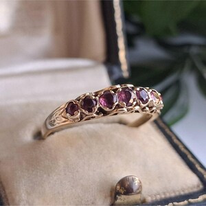 May include: A gold ring featuring six round, deep red gemstones set in a decorative band. The ring is presented in a vintage black jewellery box with a cream interior. The ring's band has intricate floral designs.