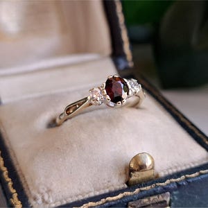 Könnte beinhalten: Ein Goldring mit einem zentralen, oval geschliffenen, dunkelroten Granat, flankiert von zwei kleinen, runden, klaren Steinen. Die Ringschiene ist nahe der Fassung geteilt und hält die drei Steine mit Metallkrappen auf einer hellen Unterlage.