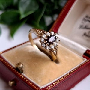 May include: A vintage gold ring with a dark red gemstone center, surrounded by smaller clear stones. The ring is displayed in a red jewelry box with a tan interior. The ring's band has an ornate design.