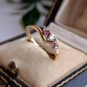 May include: A gold ring with a flowing design, featuring three pink gemstones and a central white gemstone, with small accent stones. The ring is presented in a vintage jewellery box with a cream interior.