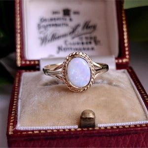 May include: A gold ring featuring an oval opal gemstone, framed by a rope-style border. The ring is presented in a red jewellery box with a cream interior. The ring's band has a split design on either side of the opal.