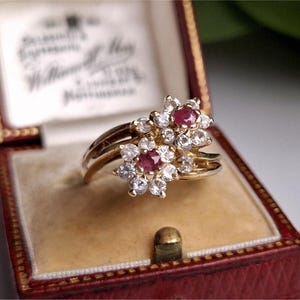 May include: A vintage gold ring with two floral clusters, each featuring a central ruby surrounded by clear gemstones. The ring is displayed in a red jewellery box with a cream-coloured interior.