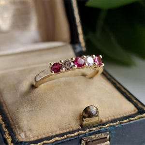 May include: A gold ring set with five gemstones. The ring has alternating red and clear stones, with a single brown stone. It is presented in a vintage blue jewellery box with a gold clasp.