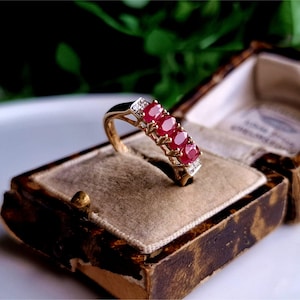 Könnte beinhalten: Ein goldener Ring mit fünf ovalen roten Edelsteinen und kleinen klaren Edelsteinen, die in die Band gefasst sind.