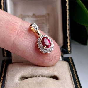 Puede incluir: Un colgante de oro con un rubí rojo central de corte ovalado rodeado por un halo de pequeños diamantes blancos redondos. El colgante presenta un engaste de borde festoneado y una pequeña anilla en la parte superior con una gema vibrante.