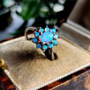 May include: A silver ring featuring an oval opal gemstone surrounded by smaller blue opal stones. The ring is set in a floral design, with the opals displaying a range of blue and iridescent colors. The ring is displayed in a vintage jewelry box.