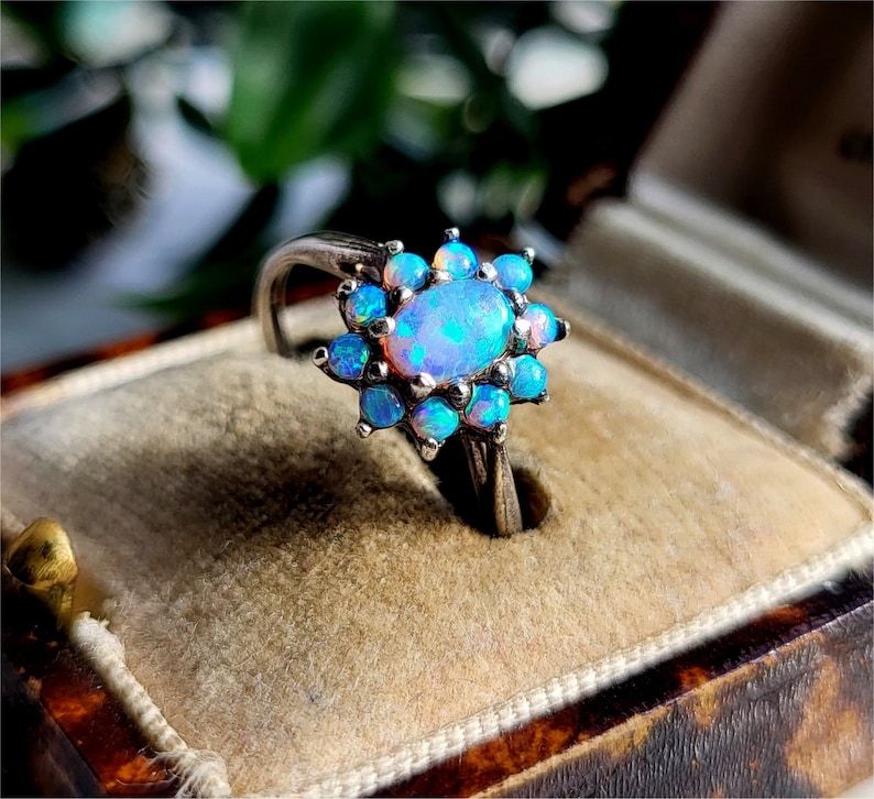 May include: A silver ring featuring an oval opal gemstone surrounded by smaller blue opal stones. The ring is set in a floral design, with the opals displaying a range of blue and iridescent colors. The ring is displayed in a vintage jewelry box.