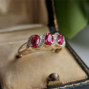 Könnte beinhalten: Ein goldener Ring mit drei ovalen, leuchtend roten Edelsteinen, die jeweils von kleinen, klaren Akzentsteinen umgeben sind. Der Ring wird in einer Vintage-Schmuckschatulle mit Samtinnenraum präsentiert.