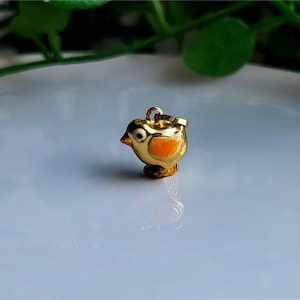 May include: A gold-coloured bird charm with black eyes, an orange beak, and orange accents on its chest and wings. The charm has a loop at the top for attaching to a necklace or bracelet. The bird is standing on a small base.