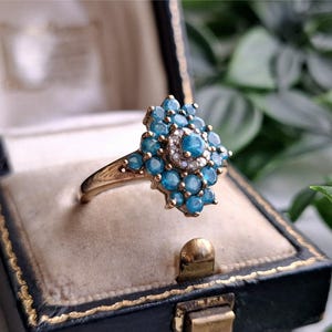 Puede incluir: Un anillo de oro con un grupo de piedras redondas de color azul brillante dispuestas en un patrón floral, con un círculo central de pequeñas piedras claras y brillantes que rodean una piedra azul en el centro.
