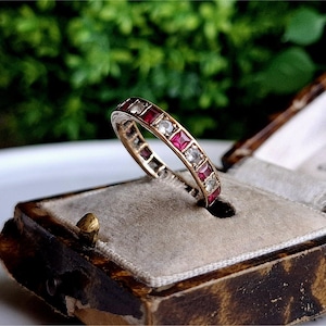 Peut inclure: Une bague en or avec des pierres précieuses rouges et blanches alternées. La bague est dans un petit coffret en velours marron.