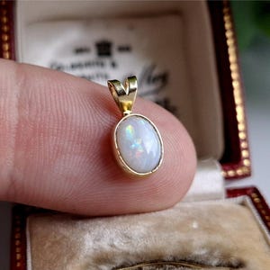 May include: A gold pendant featuring an oval opal gemstone. The opal has iridescent colours within a gold bezel setting. The pendant is held in a red jewellery box with a cream interior.