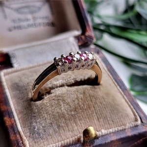 Puede incluir: Un anillo de banda de oro con cinco piedras preciosas rojas y blancas engastadas en fila. El anillo está en una caja de anillo de terciopelo marrón.