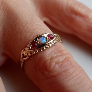 May include: A gold ring featuring an oval design with an opal center stone and two red gemstones on either side. The ring is set on a finger, showcasing its intricate details and craftsmanship. The ring's band has a textured, ornate design.
