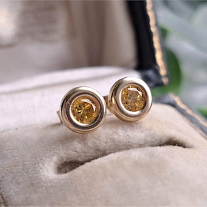 May include: A pair of stud earrings featuring round, faceted yellow gemstones set in polished gold-toned metal bezels. The earrings have a circular design with a smooth, metallic border surrounding each vibrant yellow stone.