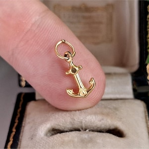 May include: A small, polished gold anchor charm featuring a classic nautical design with a rounded top loop for attachment. The charm has a smooth, reflective metallic surface and a detailed fluke structure at the base, presented in a compact size.