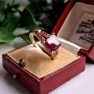 May include: A gold ring featuring a large, square, deep red gemstone and smaller red gemstones. The ring is presented in an open, red jewellery box with a gold interior. The box is lined with cream-coloured velvet.