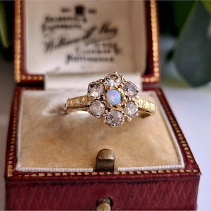 May include: A gold ring featuring a central opal stone surrounded by clear gemstones. The ring is displayed in a vintage red jewellery box with a cream interior. The ring's band has a textured design.