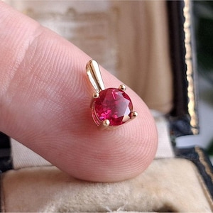 Peut inclure: Un pendentif en or avec une pierre précieuse rouge vif et ronde. Le pendentif est tenu par un doigt, mettant en valeur la couleur de la pierre et le sertissage en or. Le pendentif est probablement un bijou.