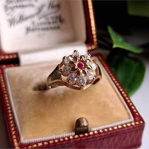 May include: A gold ring with a floral design, featuring a central red gemstone surrounded by clear, round-cut stones. The ring is displayed in a red and gold jewelry box with a cream-coloured interior. The box is open, revealing the ring.
