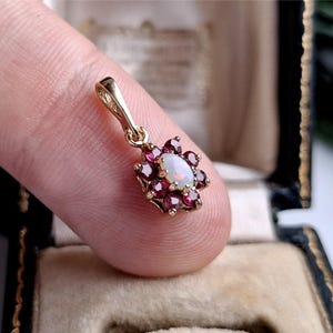 May include: A gold pendant featuring a central oval-shaped white opal surrounded by a cluster of ten small, deep red faceted garnet stones. The pendant has a polished gold bail at the top for attachment to a chain, showcasing a floral-like design.