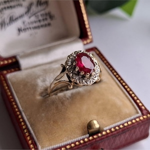 Może przedstawiać: Złoty pierścionek z dużym owalnym rubinem w środku, otoczonym mniejszymi, przezroczystymi kamieniami. Pierścionek jest prezentowany w czerwonym pudełku na biżuterię z kremowym wnętrzem.