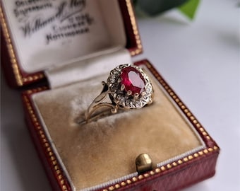 Könnte beinhalten: Ein goldener Ring mit einem großen ovalen roten Mittelstein, umgeben von kleineren klaren Steinen. Der Ring wird in einer roten Schmuckschatulle mit cremefarbenem Innenraum präsentiert.