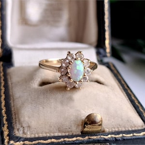 May include: A gold ring featuring a central oval-shaped opal gemstone surrounded by a halo of ten small, clear round-cut stones. The ring is set in a polished gold band, showcasing a classic cluster design with a vibrant, iridescent opal centre.