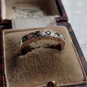 Puede incluir: Un anillo de banda de oro con un diseño delicado que presenta piedras preciosas verdes y blancas alternas. El anillo se muestra en una caja forrada de terciopelo.