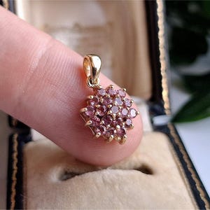 Peut inclure: Un pendentif en or orné d'un amas de petites pierres précieuses roses et rondes. Le pendentif est tenu entre les doigts, mettant en valeur son design complexe et l'éclat des pierres précieuses. Le sertissage semble être en or.