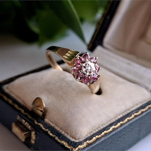 Può includere: Un anello in oro con montatura a grappolo caratterizzato da una pietra centrale trasparente circondata da otto piccole pietre preziose rotonde di colore rosa intenso. La fascia presenta un design a coste testurizzato ed è in un cofanetto blu.