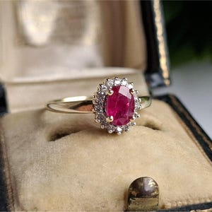 May include: A gold ring with a large oval ruby center stone, surrounded by a halo of small diamonds. The ring is set in a vintage-style setting and is displayed in a velvet-lined jewelry box.