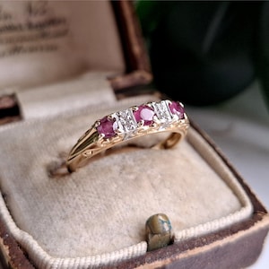 Könnte beinhalten: Ein Goldring mit drei runden, roten Rubinen, die sich mit zwei kleinen, klaren Steinen in einer Reihe abwechseln. Der Ring hat eine polierte Oberfläche und dekorative Metallarbeiten an den Seiten sowie eine sichere Krappenfassung.