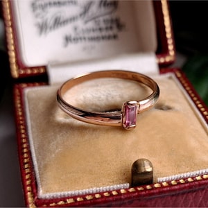 Könnte beinhalten: Ein goldener Ring mit einem rechteckigen rosa Edelstein, präsentiert in einer roten Schmuckschatulle mit cremefarbenem Innenraum. Der Ring hat ein schlichtes Banddesign. Die Schatulle ist geöffnet und zeigt die Details des Rings.