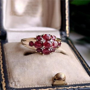May include: A gold ring featuring a cluster of oval-cut red rubies arranged in a floral pattern, accented with small clear stones set between the rubies. The band is smooth and polished gold, showcasing a classic design with multiple gemstones.