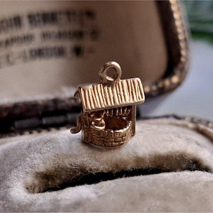 May include: A gold-coloured well charm, featuring a small roof and a bucket detail. The well is intricately designed with brickwork and a loop for attachment. The charm is displayed on a soft, velvet surface, suggesting a jewellery setting.