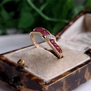 May include: A gold band ring with a row of red rubies and a single diamond set in the center. The ring is in a small, brown velvet box.