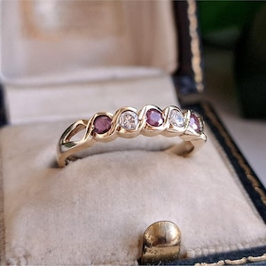 Könnte beinhalten: Ein Goldring mit einer abwechselnden Reihe aus drei runden roten Edelsteinen und zwei runden klaren Edelsteinen, die in ein gedrehtes Metallbanddesign eingefasst sind. Der Ring wird in einer dunklen Schmuckschatulle präsentiert.