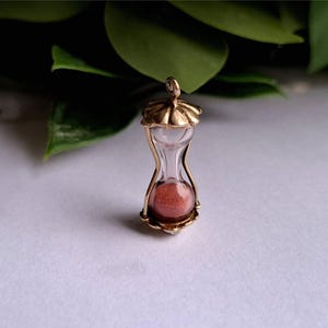 Può includere: Un piccolo ciondolo a forma di clessidra con una montatura color oro e un centro in vetro trasparente. La clessidra contiene sabbia rossa. La parte superiore e inferiore del ciondolo hanno un design decorativo smerlato. Il ciondolo è probabilmente in metallo.