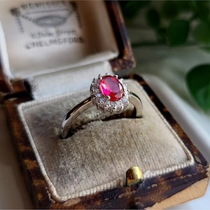 Könnte beinhalten: Ein silberner Ring mit einem roten ovalen Edelstein, der von kleineren weißen Edelsteinen umgeben ist. Der Ring befindet sich in einer braunen Samt-Ringbox.