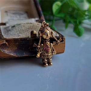 Könnte beinhalten: Ein goldfarbener Anhänger in Form einer Puppe mit rosafarbenen und weißen Edelsteinen für die Augen und einem rosafarbenen Edelstein-Mieder. Die Puppe hat eine goldfarbene Kette, die an der Oberseite ihres Kopfes befestigt ist.
