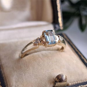 May include: A gold ring featuring a rectangular, light blue gemstone. The ring has a detailed design with small accent stones flanking the main stone. It is presented in a vintage jewellery box with a cream-coloured interior.