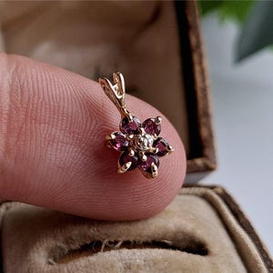May include: A gold pendant in the shape of a flower, with dark red gemstones and a central diamond. The pendant is held by a finger, with a jewelry box in the background. The flower has six petals and a small loop for a chain.