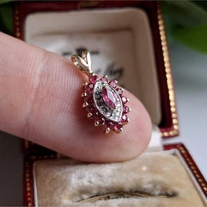 May include: A gold pendant with a central marquise-cut pink gemstone, surrounded by smaller red gemstones and a halo of white stones. The pendant is set in a gold frame and has a loop for a chain.