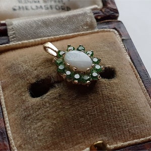 Puede incluir: Colgante de oro con una ópalo ovalada central rodeada de pequeñas piedras verdes. El colgante está engastado en un marco de oro y se muestra en un joyero vintage marrón con interior beige.