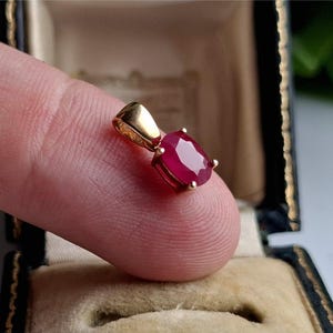 Puede incluir: Un colgante de oro con una vibrante gema roja de corte ovalado. El colgante está sostenido por un dedo, mostrando el rico color de la gema y el brillo del engaste de oro. El colgante se exhibe en un joyero vintage.