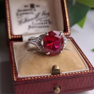 May include: A silver ring with a large, round, red gemstone is presented in a red jewellery box with a beige interior. The ring's setting has decorative accents. The box is open, revealing the ring and the interior lining.