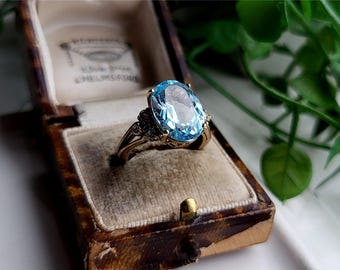 May include: A gold ring with a large oval-shaped blue topaz gemstone and smaller diamond accents. The ring is in a brown velvet ring box.