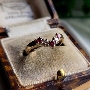 May include: A gold ring with a curved design, set with small red gemstones and a few clear stones. The ring is displayed inside a vintage, patterned jewellery box with a velvet interior.