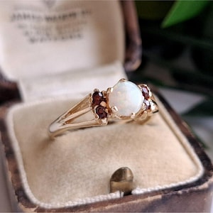 May include: A gold ring featuring a central round cabochon opal stone flanked by two small dark red garnet stones on each side. The ring has a split shank design and is presented in a beige velvet-lined jewelry box with a dark brown exterior.